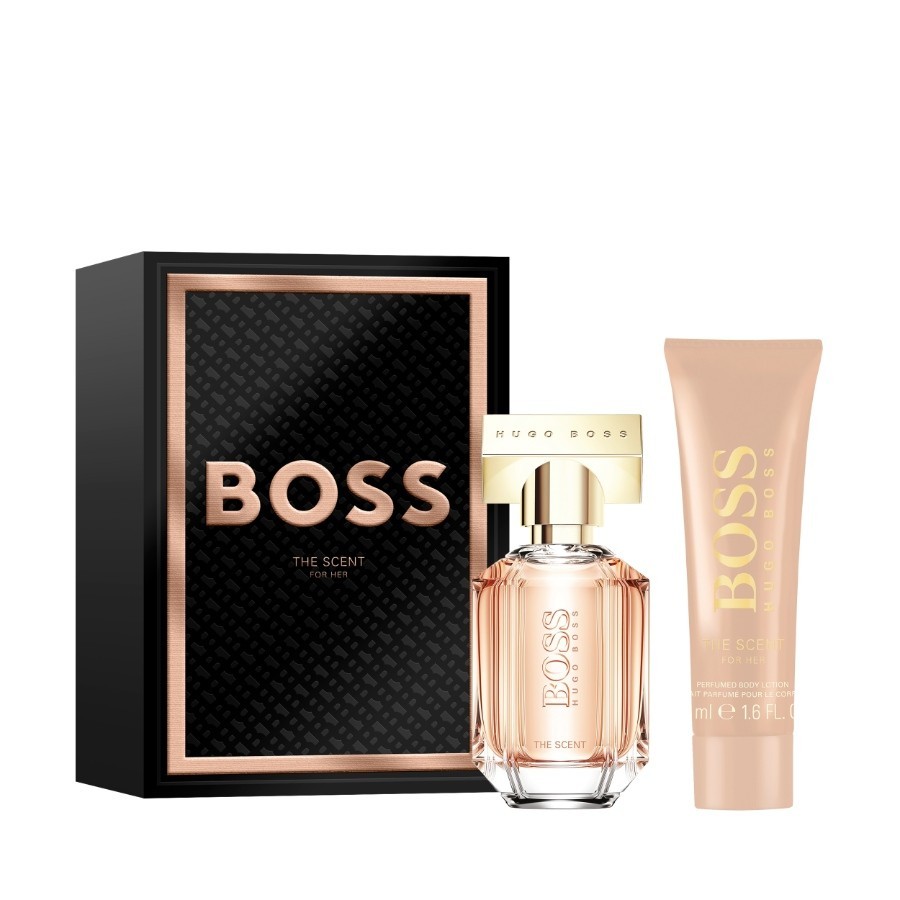 Black gift box with gold accents featuring "BOSS THE SCENT" label, alongside a clear glass perfume bottle and a matching cream tube, both in soft pink hues.