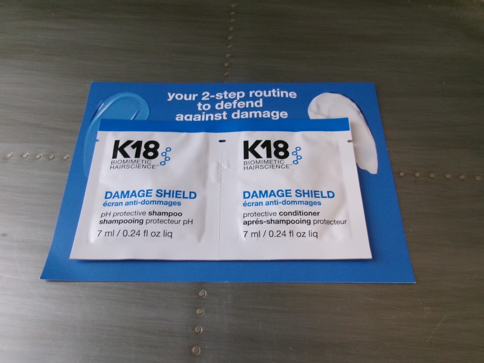 Two white sachets labeled "K18 Damage Shield" for shampoo and conditioner, featuring a blue background with text about a two-step routine to defend against damage.