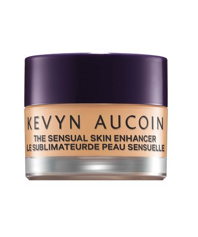 A small, round jar of Kevyn Aucoin The Sensual Skin Enhancer, featuring a purple lid and a clear base, with a light beige product visible inside.
