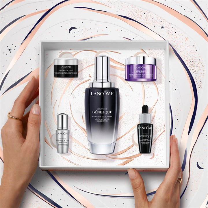 A gift set featuring Lancôme's Advanced Génifique Serum in a sleek silver and black bottle, alongside a purple cream and two smaller serums, all presented in an elegant box with a swirled design.