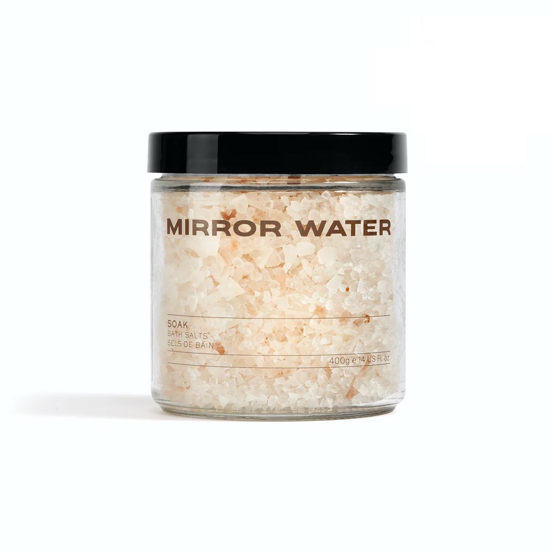 Clear glass jar filled with coarse bath salts in shades of white and light pink, featuring a black lid and a label that reads "MIRROR WATER Soak" in bold, brown lettering.