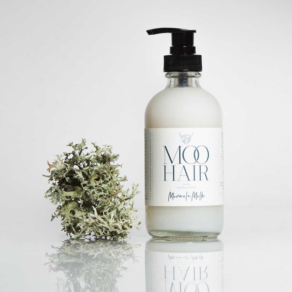Clear glass pump bottle of "Miracle Milk" by Moo Hair, featuring a white label with blue text, alongside a decorative piece of moss.