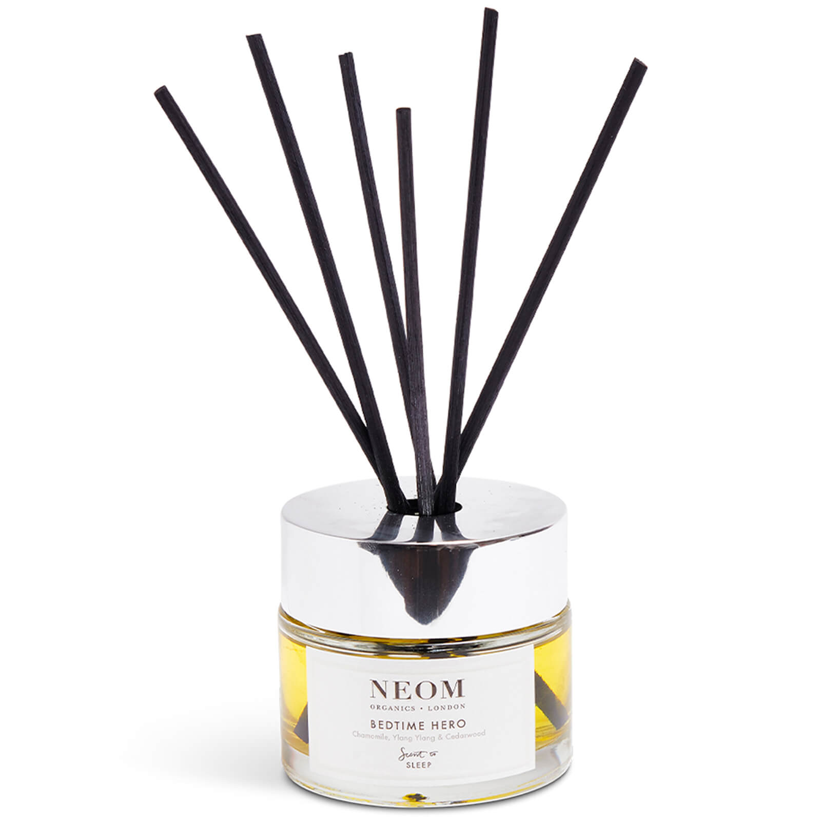 Clear glass diffuser with a silver lid, containing yellow liquid and six black reed sticks, labeled "NEOM Bedtime Hero" in elegant font.