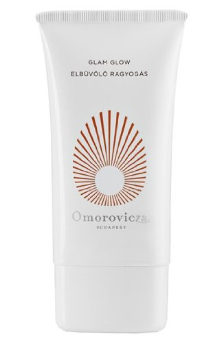 White tube with a sleek design featuring a sunburst pattern in copper, labeled "Glam Glow" by Omorovicza, with a rounded cap and elegant branding.