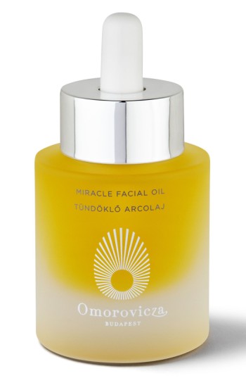 A frosted glass bottle with a silver dropper cap, containing yellow facial oil, labeled "Miracle Facial Oil" with the Omorovicza logo and a sunburst design.