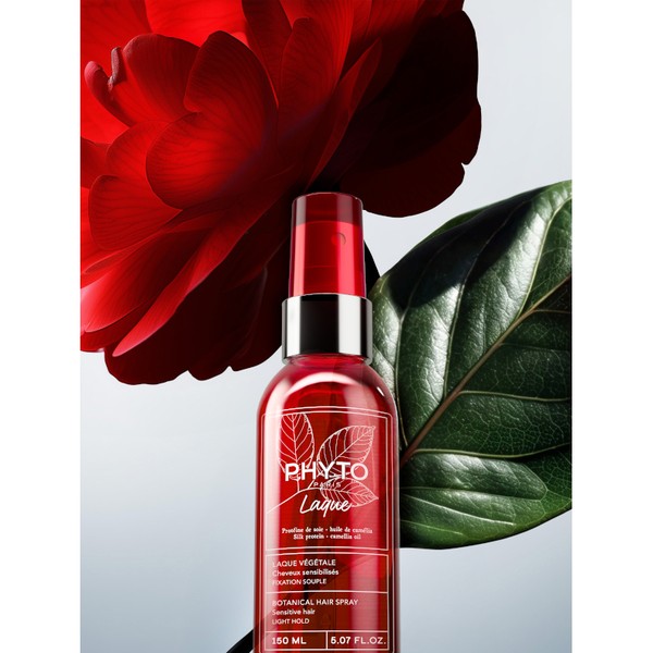 Transparent spray bottle with a red cap, featuring a botanical design, labeled "Phyto Laque Flexible Hold Hairspray," set against a backdrop of a large red flower and green leaves.