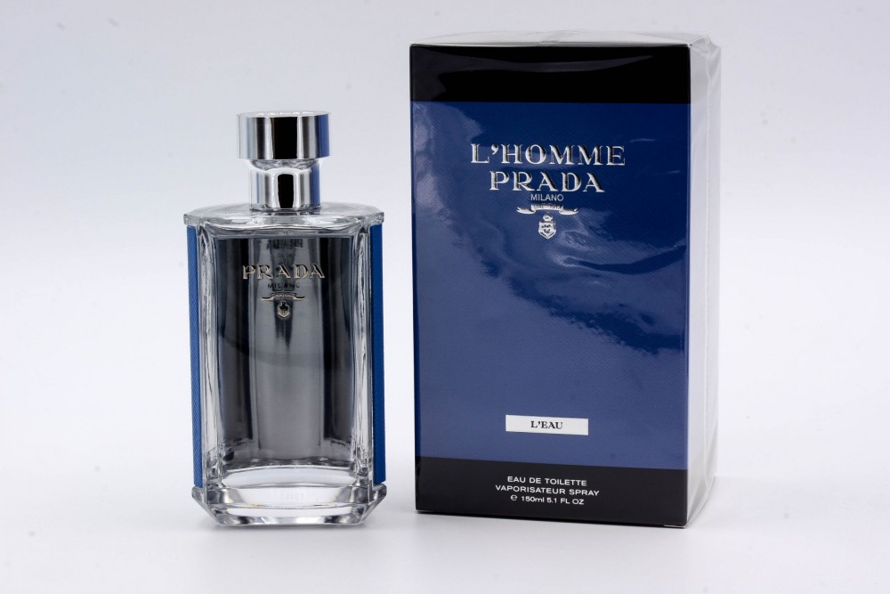 Clear glass bottle with a silver cap, featuring a sleek design and engraved "Prada" logo, alongside a blue box labeled "L'Homme Prada Milano L'Eau."