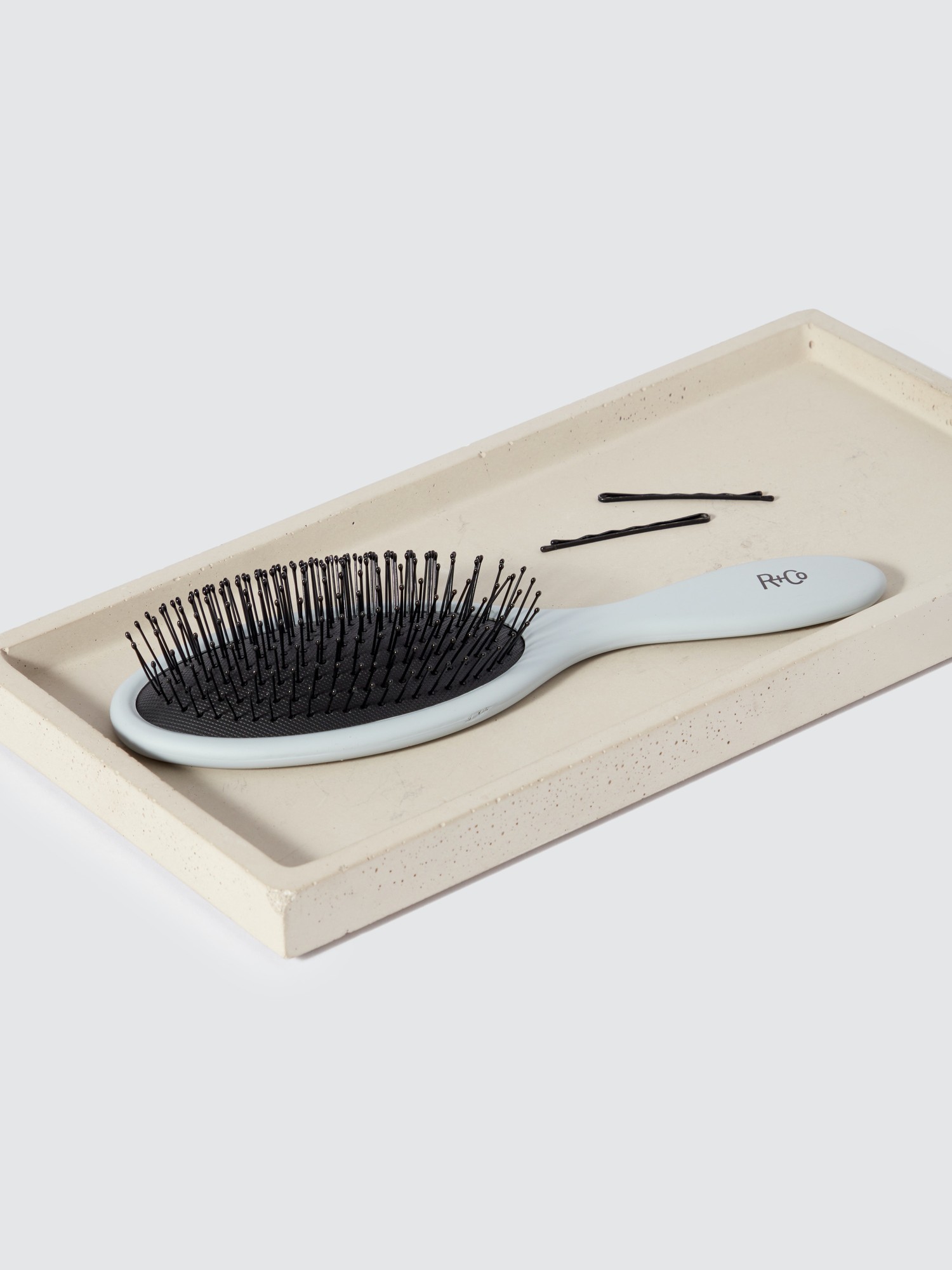 A white and black detangling brush with flexible bristles, resting on a light-colored tray, accompanied by a few bobby pins.