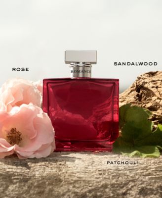 Ralph Lauren Romance Eau De Parfum Intense in a deep red glass bottle, featuring a silver cap, surrounded by pink roses and green leaves, highlighting notes of rose, sandalwood, and patchouli.