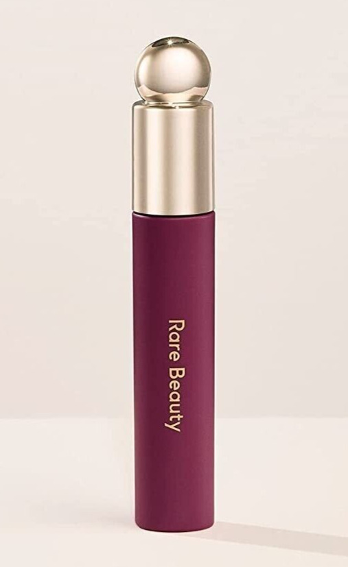 Deep burgundy lip oil stain in a sleek tube with a gold cap, featuring "Rare Beauty" embossed in gold on the side, designed for a soft, tinted finish.