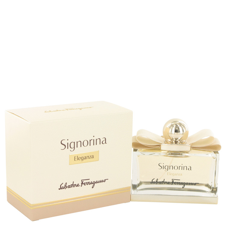 Clear glass bottle with a round gold cap, adorned with a delicate beige ribbon, next to a cream-colored box featuring elegant gold lettering for "Signorina Eleganza" by Salvatore Ferragamo.