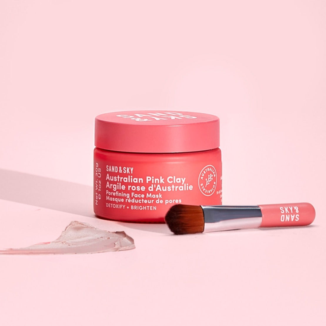 Australian Pink Clay Porefining Face Mask in a pink jar with a white label, accompanied by a small brush and a swatch of the clay on a light pink background.