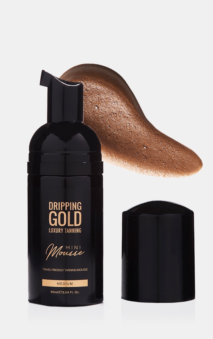 Black pump bottle with gold accents, containing 90ml of medium self-tanning foam, with a rich brown mousse texture spilling out, designed for travel use.