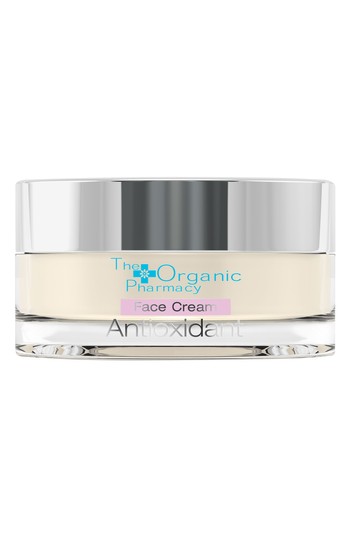 Clear glass jar with a silver lid, containing a creamy, pale yellow face cream labeled "Antioxidant" and "The Organic Pharmacy" in light blue and pink text.