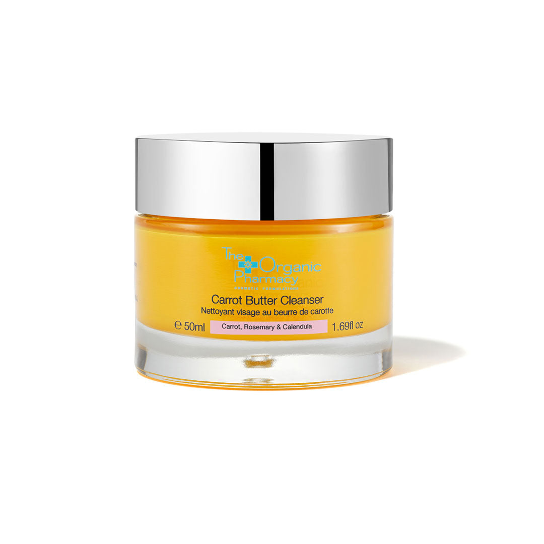 A clear glass jar with a silver lid contains a bright orange carrot butter cleanser labeled "Carrot Butter Cleanser" by The Organic Pharmacy, 1.7 oz.