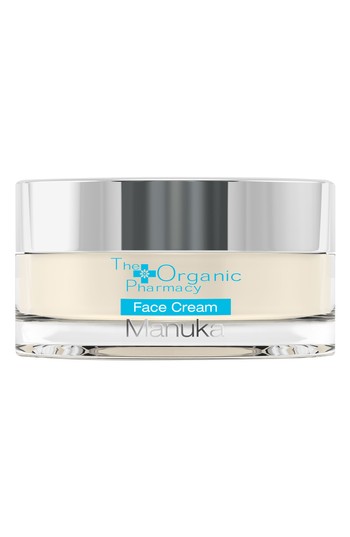 A clear glass jar with a silver lid, containing a creamy beige face cream labeled "The Organic Pharmacy Manuka Face Cream" in blue text.