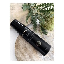Black bottle of Three Warriors Self-Tan Mousse with a sleek design, featuring a silver logo and cap, placed on a light wooden surface surrounded by green foliage.
