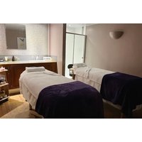 Two massage tables with white linens and purple blankets in a softly lit spa room with a wooden cabinet and mirror.