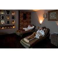 Two people in white robes relaxing on loungers in a dimly lit spa room with candles and soft lighting.