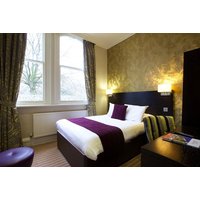 Hotel room with double bed, purple accents, patterned curtains, and a view of trees through large windows.
