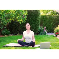 Woman meditating on a pink mat in a garden with a laptop nearby, promoting a remote guided meditation session.