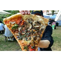 Large slice of pizza with mushrooms, spinach, and tomatoes, held up outdoors in a park setting.