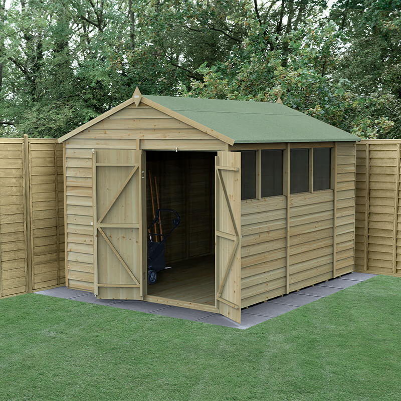 A wooden apex shed with a green felt roof, featuring double doors, large windows, and pressure-treated timber, set against a backdrop of trees and a wooden fence.