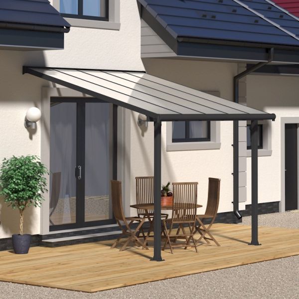 Grey patio cover with clear panels, supported by black metal posts, extending over a wooden deck with a round table and chairs, adjacent to a house with a modern design.