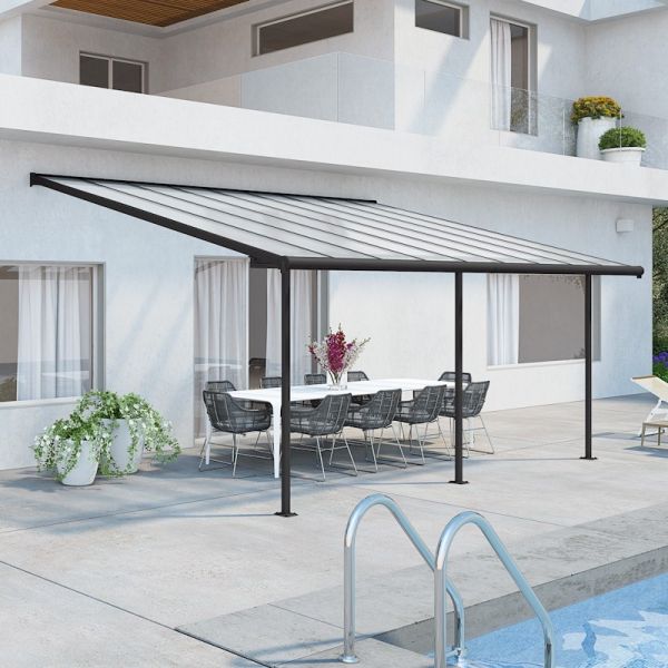 A grey patio cover with a slanted roof, supported by black metal posts, provides shade over a dining table and chairs on a stone patio near a pool.