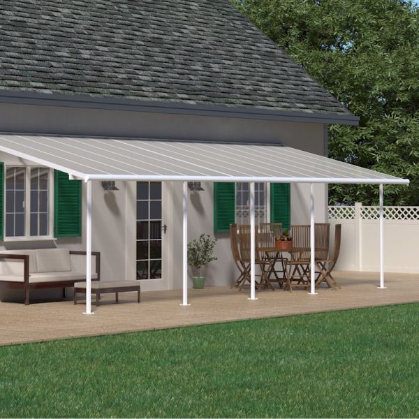 White 10'x30' Palram Canopia Sierra patio cover with a slanted roof, supported by white posts, shading a wooden deck with outdoor furniture and green shutters on a house.
