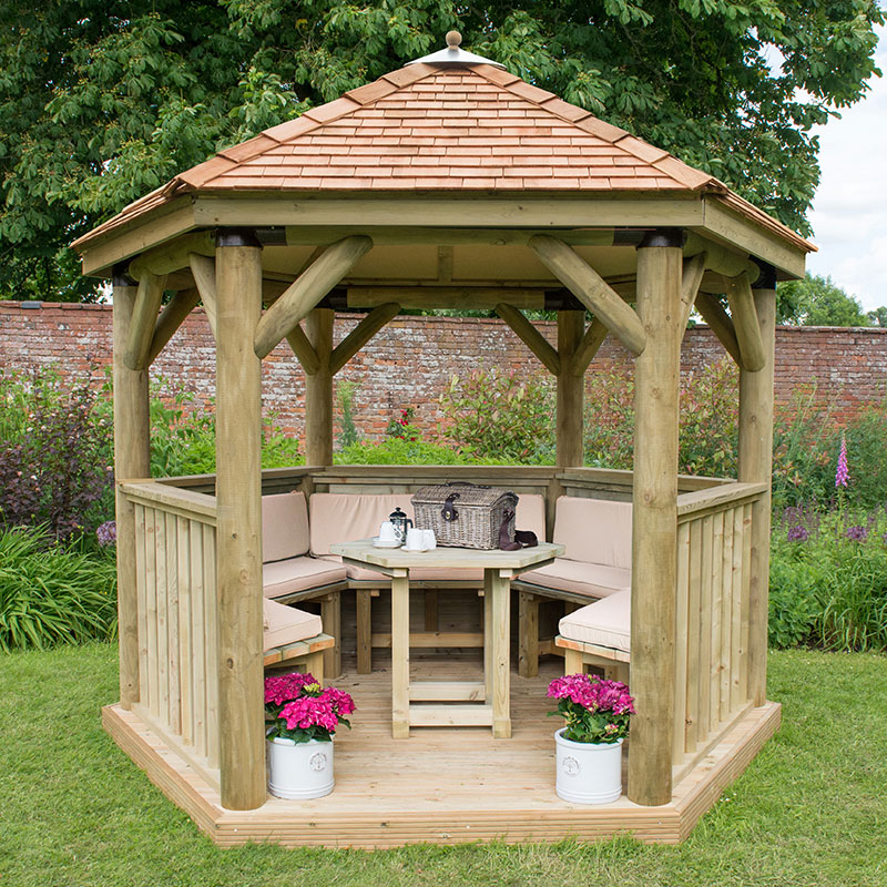 Luxury wooden gazebo with a New England cedar roof, featuring light-colored wooden beams, cushioned seating for up to 10, and flower pots at the base, set in a lush garden.