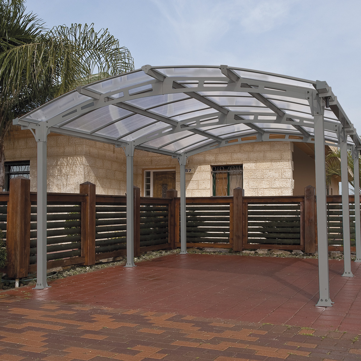 Grey metal carport with a curved roof and transparent panels, supported by sturdy posts, set on a brick surface with wooden fencing in the background.