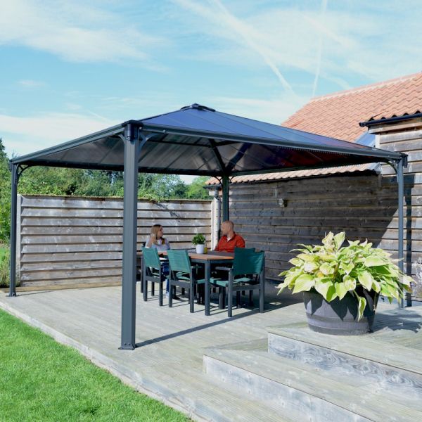 Grey 14'x14' Palram Canopia Palermo gazebo with a sturdy aluminum frame, polycarbonate roof, and a wooden deck, featuring two people seated at a dining table under the structure.