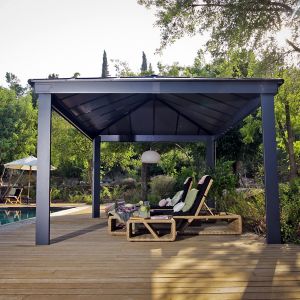 A grey, 16' x 12' Palram Canopia Dallas gazebo with a slanted roof, supported by sturdy black posts, features lounge chairs on a wooden deck beside a pool.