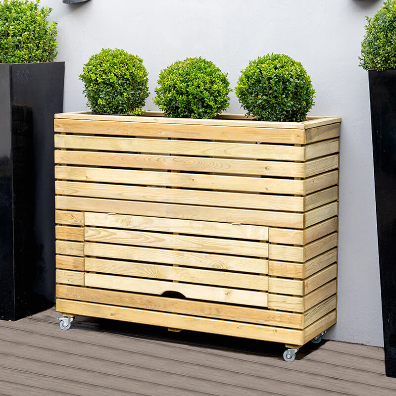 Tall wooden garden planter in natural wood finish, featuring horizontal slats, a storage compartment, and wheels for mobility, topped with three round green topiary plants.
