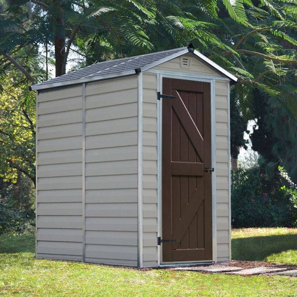 Tan plastic shed with a sloped roof, featuring a brown double door, vertical paneling, and a skylight, set in a green outdoor environment with trees in the background.