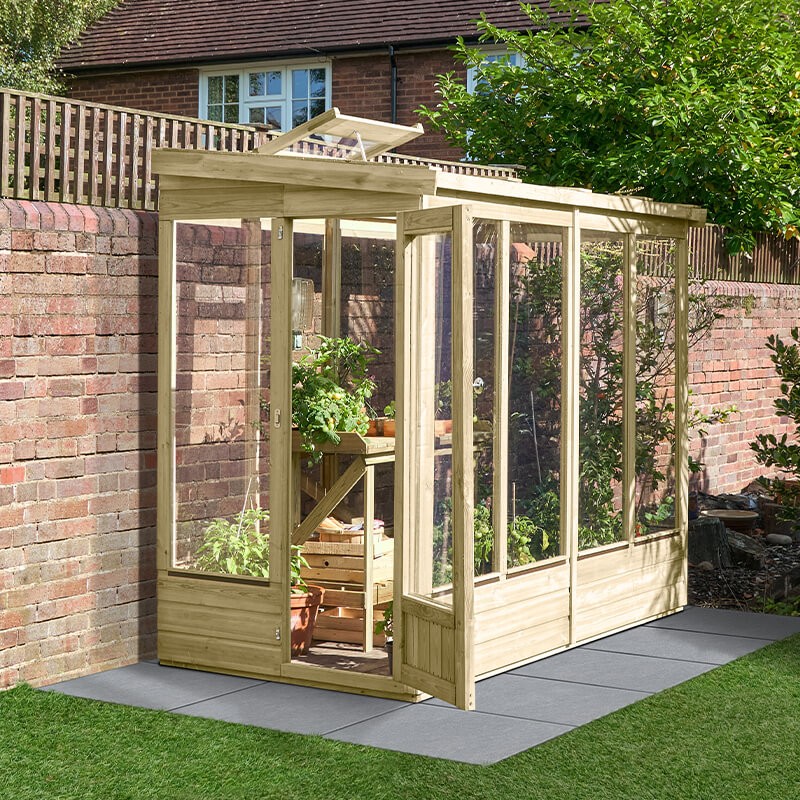 A wooden lean-to greenhouse with a glazed back, featuring large glass panels, a slanted roof, and a wooden door, set against a brick wall and surrounded by greenery.