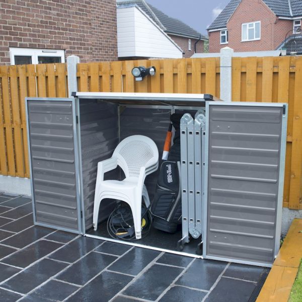 Dark grey Palram Canopia Voyager shed with double doors, featuring a sturdy metal design, housing a white chair, folding table, and various tools on a paved surface.