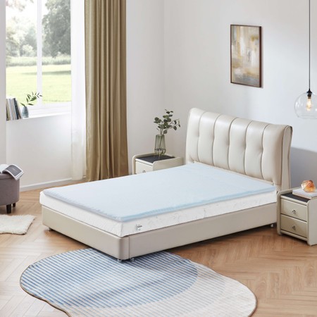 5-zone memory foam mattress topper in light blue with a soft cover, placed on a beige upholstered bed in a bright room with wooden flooring and natural light.