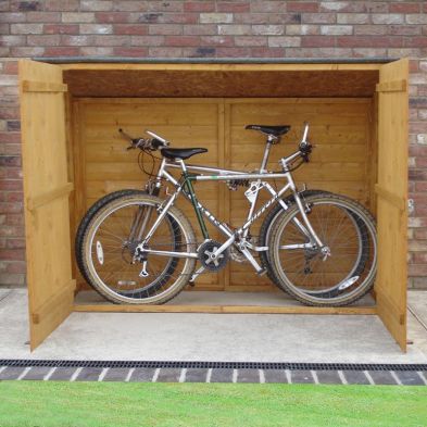 Wooden bike shed in natural finish, measuring 6' x 2'6", featuring double doors and no floor, with two bicycles stored inside against a brick wall.