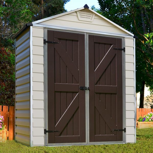Tan plastic shed with a peaked roof, featuring double brown doors with diagonal slats, metal hinges, and a skylight, set against a green lawn and garden backdrop.
