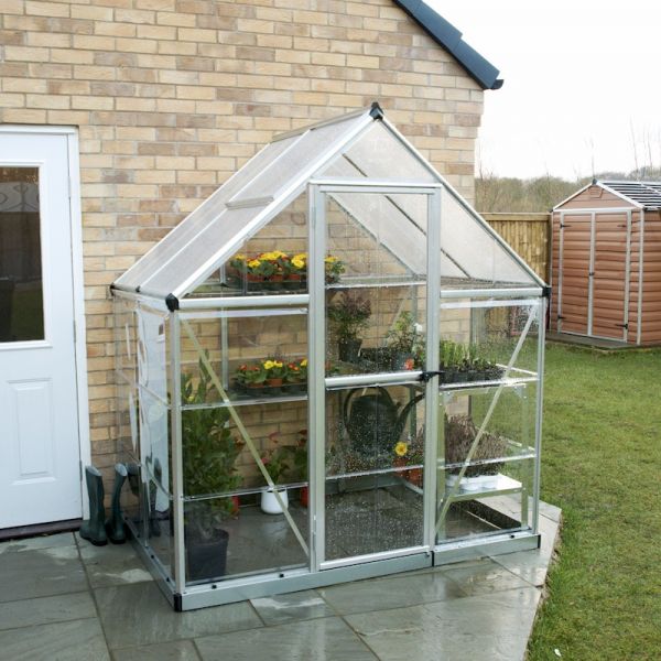 Silver polycarbonate greenhouse with a sloped roof, featuring clear panels, a double door, and shelves, situated beside a brick wall and green lawn.