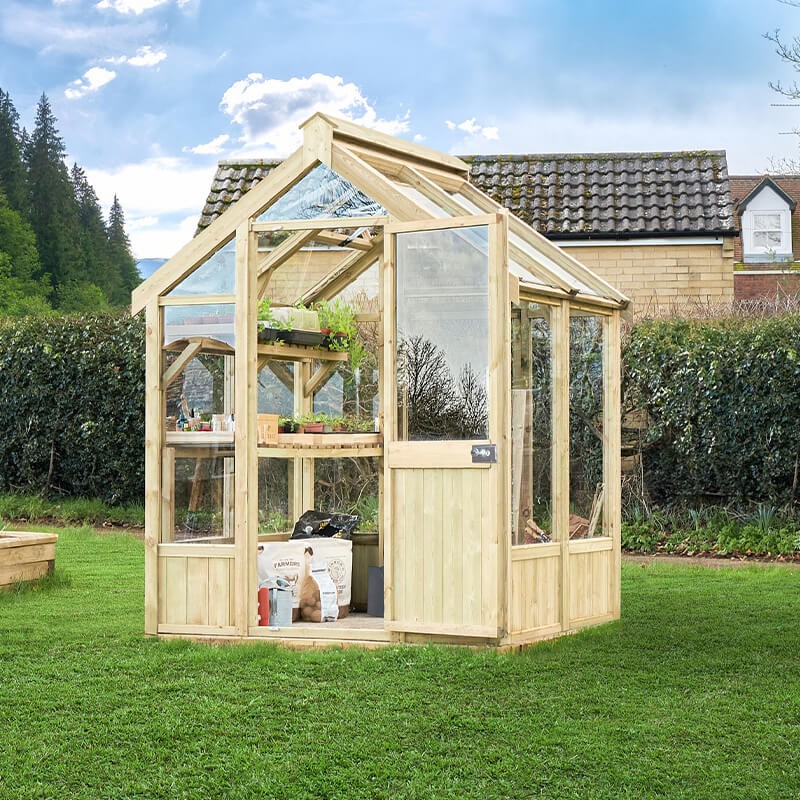 A wooden greenhouse with a natural finish, featuring large glass panels, a sloped roof, and a door, set in a grassy area with trees and a house in the background.