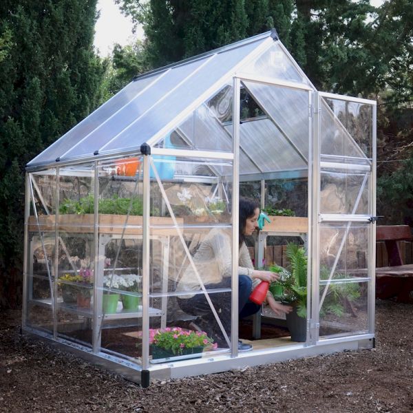 A silver polycarbonate greenhouse with a sloped roof, featuring transparent panels, wooden shelves filled with plants, and a woman tending to a potted fern inside.