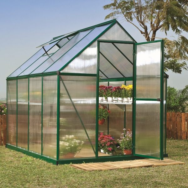 Green polycarbonate greenhouse with a walk-in design, featuring a double door, slanted roof, and plants on shelves inside, set on a grassy area with wooden fencing in the background.