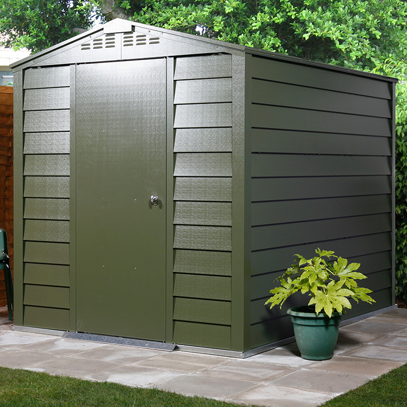 Green metal shed with a sloped roof, vertical paneling, and a single door, set on a paved area with a potted plant nearby and surrounded by greenery.