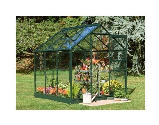 Small greenhouse featuring a green frame, glass panels, and a sloped roof, surrounded by colorful flowers and plants on a grassy area.