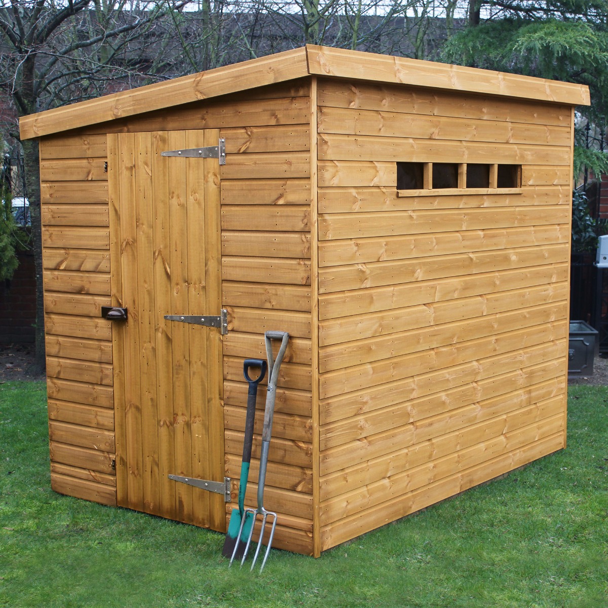 Traditional wooden garden shed in a warm brown finish, featuring shiplap siding, a pent roof, a single door with a latch, and a small window, set on a grassy area with garden tools nearby.