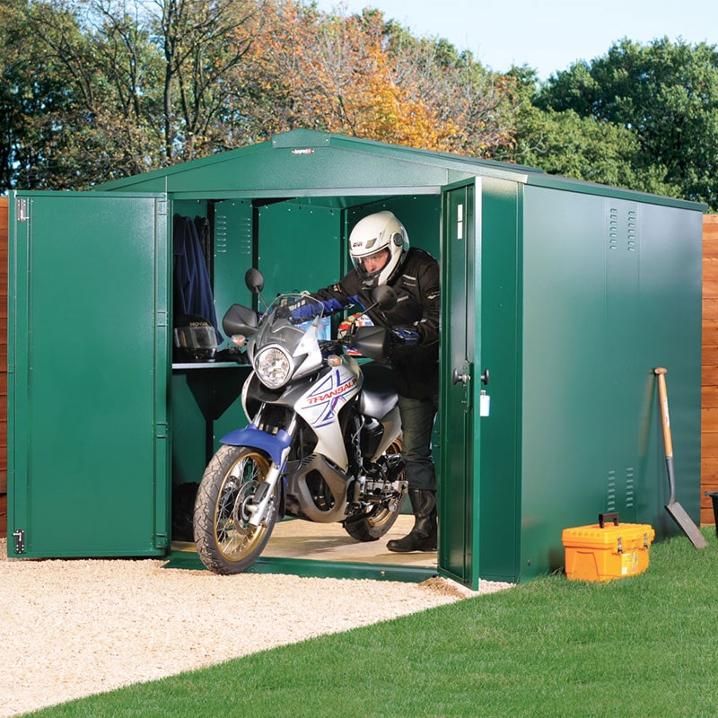 Green metal garage with a sloped roof, featuring double doors, housing a motorbike and a rider in protective gear, set on a gravel surface with trees in the background.
