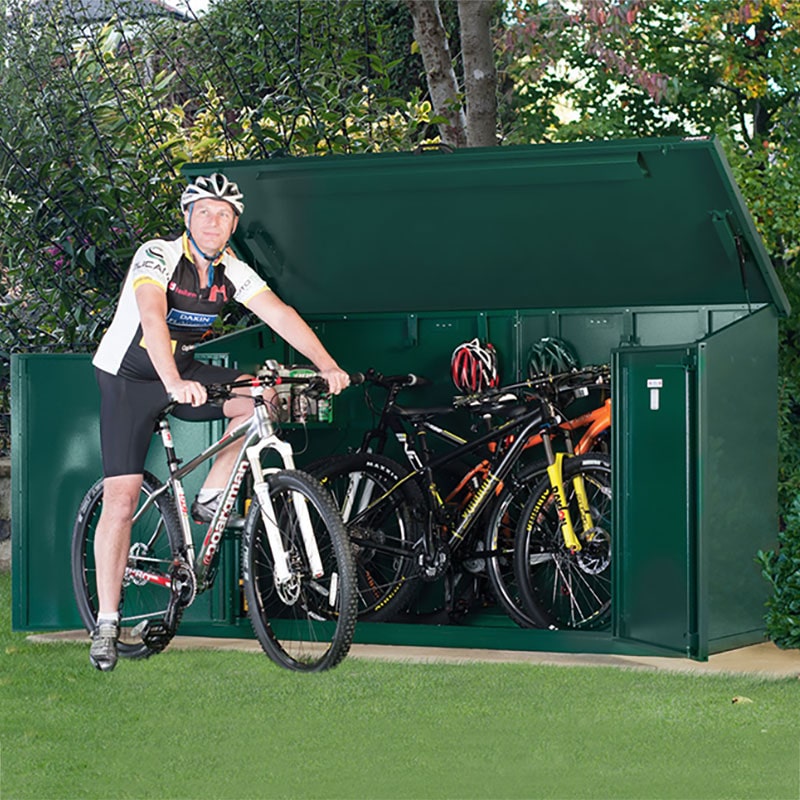 Green metal bike shed with a sloped roof, designed to hold four bikes, featuring a cyclist standing next to it, showcasing ample storage space and secure access.